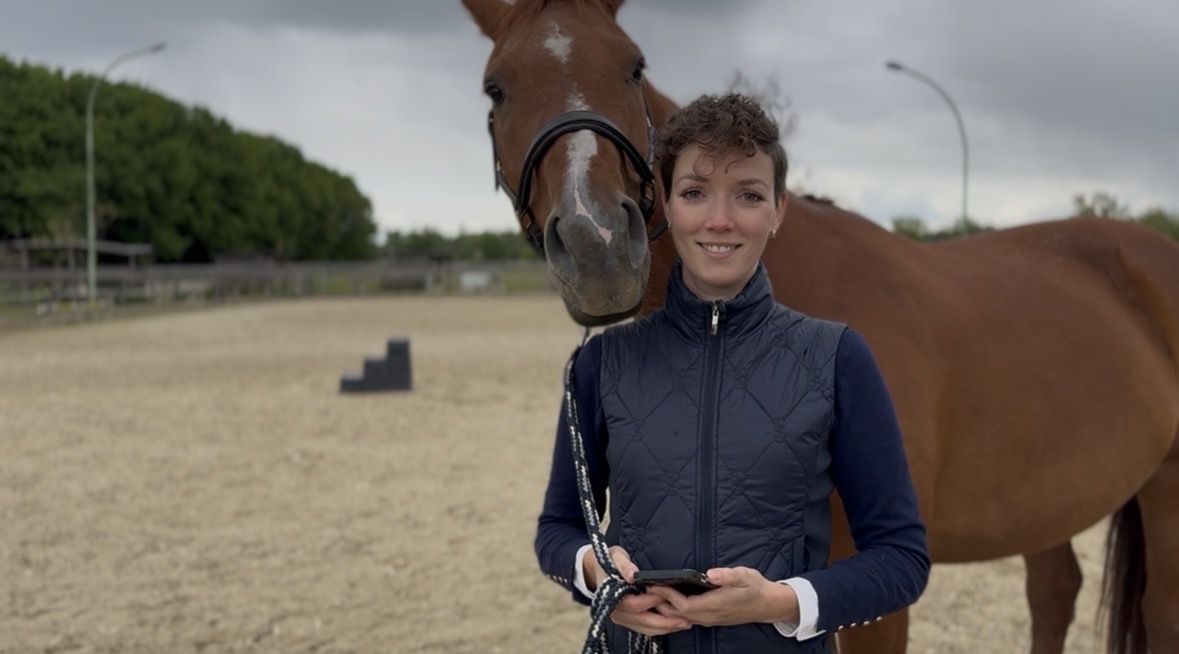 Pferdebesitzerin mit Smartphone steht neben ihrem Pferd auf dem Reitplatz